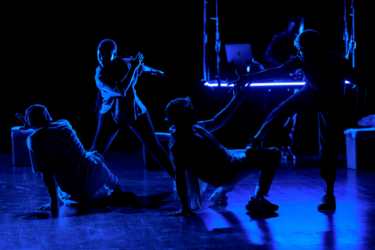A group of five dancers from Via Katlehong dance company performing on stage, almost completely in shadow with their outlines illuminated in blue light.