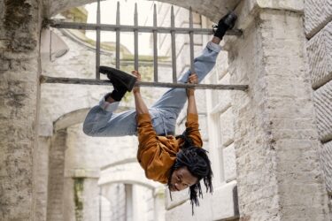 Evion Hackett hanging upside down from railings beneath a stone arch at Somerset House.