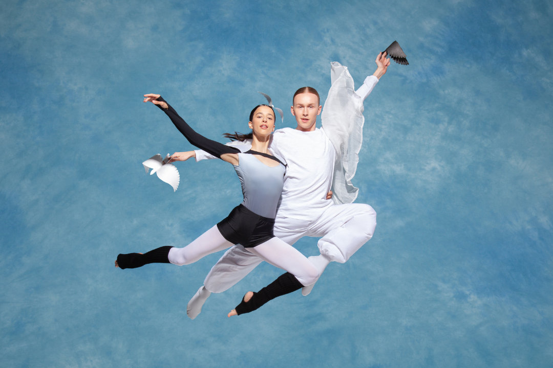 Two dancers leaping in mid-air against a sky blue backdrop. They are each holding a paper bird.