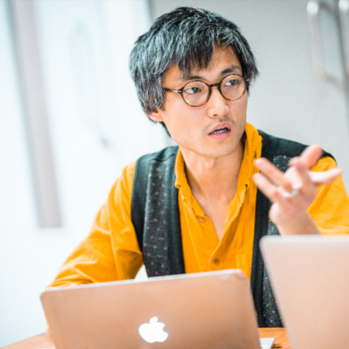 Portrait of Sho Shibata, wearing a mustard yellow shirt and grey knit open vest, gesticulating in front of a Mac laptop