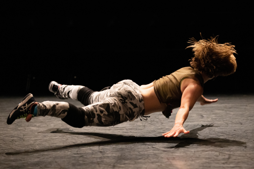 A dancer falling face down with their arms and legs spread out, hovering just above the stage.