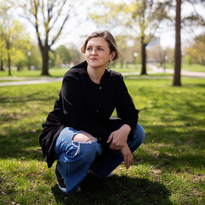 Abby Zbikowski wearing a black top and ripped blue jeans, crouching low with her arms rested on her knees. She is in a park.