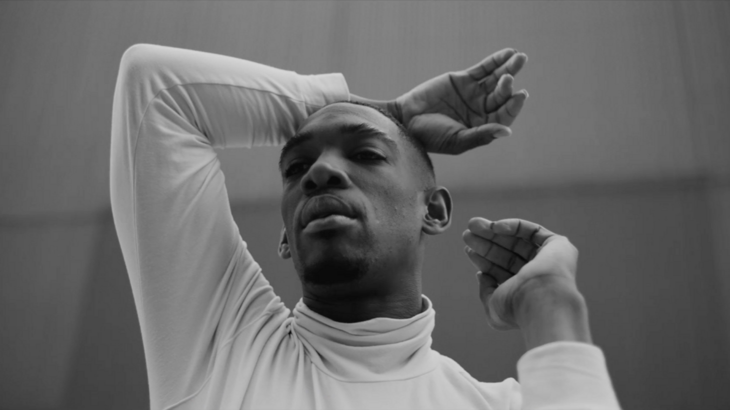 Black and white still from the film The Language of Jay Jay Revlon, wearing a white turtleneck, with his right arm raised.