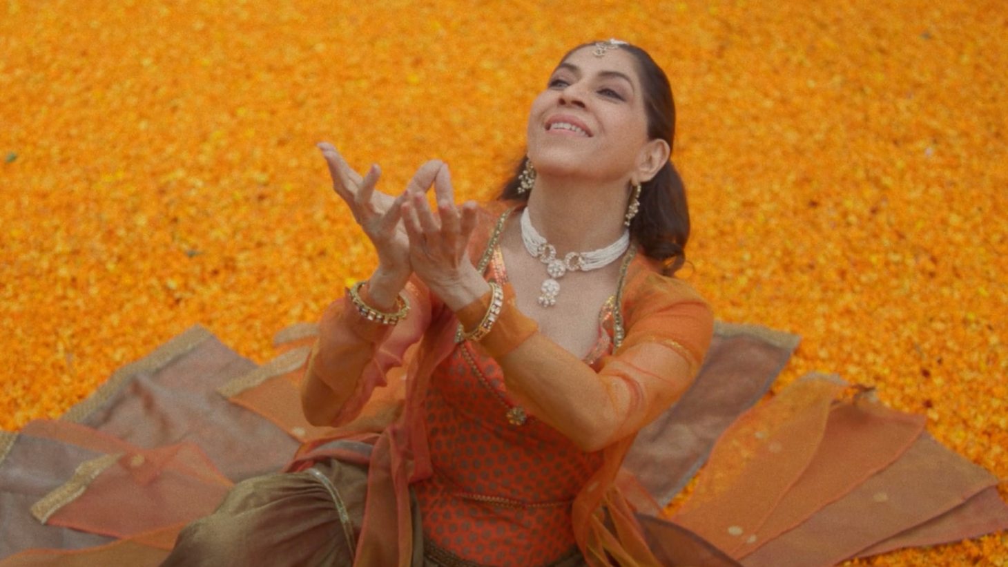 Still from the film Widening Circles by Aditi Mangaldas. Aditi is sitting on a floor of orange marigold petals, wearing an orange sari. She is looking upwards, with both hands reaching upwards.