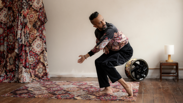 Hetain Patel wears a bomber jacket in the same geometric pattern as the rug beneath him and the drapes behind him. He is barefoot, legs bent, crouching forward, with his head turned to look behind him.