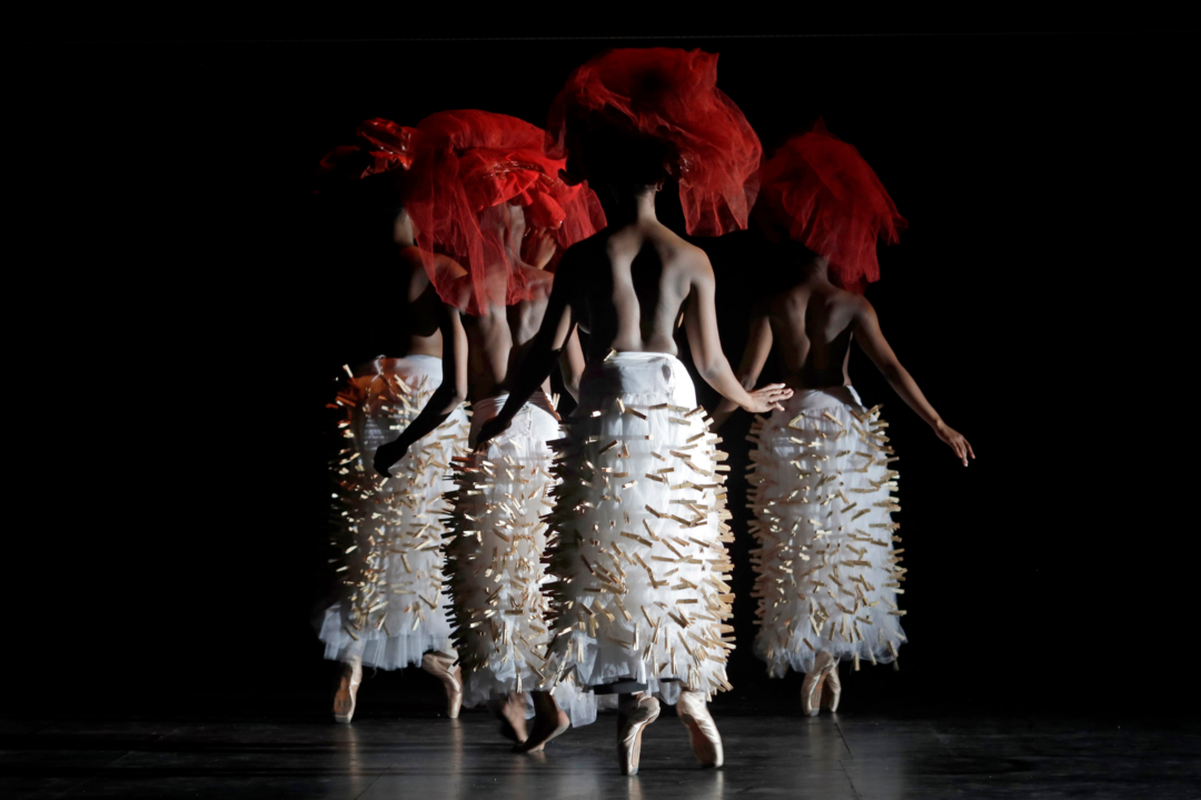 Four dancers en pointe with their backs to the camera. They are wearing long white tutu skirts with pegs attached and large red net head pieces.