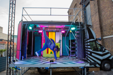 Photo of The Bobby Dazzler. A black and white striped van has the side panel removed to transform the van into a performance stage. The inside of the van is covered in colourful lit-up panels. A dancer in a blue tracksuit is doing a hand stand one hand, with both legs in the air.