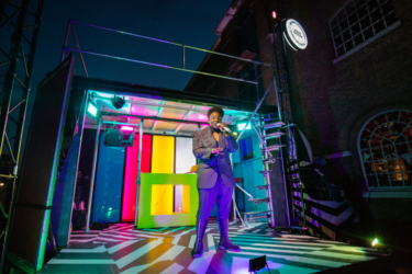 Photo of The Bobby Dazzler. A black and white striped van has the side panel removed to transform the van into a performance stage. The inside of the van is covered in colourful lit-up panels. A figure wearing dressed in a bedazzled suit is talking into a microphone.