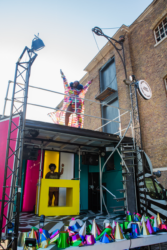 Photo of The Bobby Dazzler. A black and white striped van has the side panel removed to transform the van into a performance stage. Inside the van there is a DJ with their left arm raised. On top of the van there is a dancer wearing a multi-coloured coat with their arms raised high in the air, smiling.