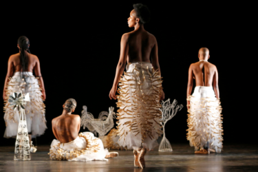 Four performers on stage with their backs to their camera, apart from one whose head is turned to the side. Three are standing, one is sitting. They are each wearing long white tutu skirts that are covered in pegs.