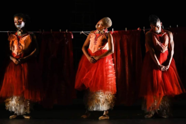 Three dances dressed in sheer red netting stand in front of a clothes line, with red netting attahced by pegs. They each have their arms in front of their bodies, one hand wrapped over the other.