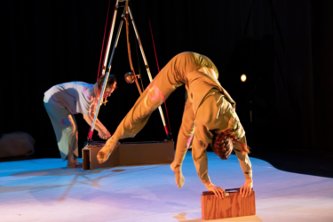 Production image from Eyecatchers. A dancer in a beige coloured boiler suit is doing a half-hand stand, with both hands holding onto a block of wood.