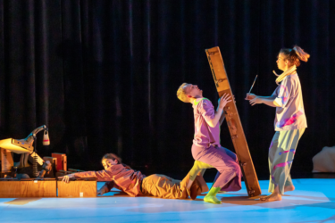 Production photo from Eyecatchers. A dancer is holding up a heavy wooden pillar, sitting on the upturned feet of another dancer who is lying on the floor. A third dancer is looking on.