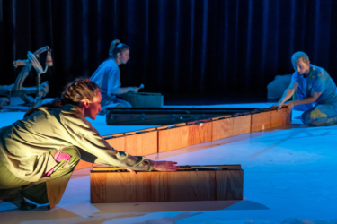 Production photo from Eyecatchers. The stage is covered in blue light. The dancer in the foreground is lit by a warm pink light, with their arms outstretched holding onto a wooden contraption. In the background there is another dancer doing the same, and further dancer who is holding a musical mallet.