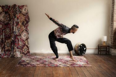 Hetain Patel wears a bomber jacket in the same geometric pattern as the rug beneath him and the drapes behind him. He is barefoot, legs bent, bent forward, with his right arm outstretched behind him.