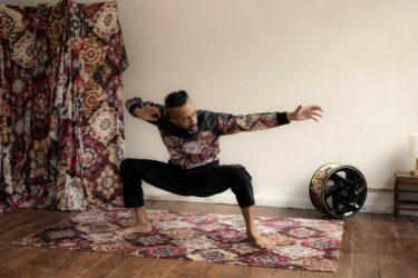 Hetain Patel wears a bomber jacket in the same geometric pattern as the rug beneath him and the drapes behind him. He is barefoot, legs crouched low, wide apart with arms outstretched.