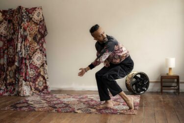 Hetain Patel wears a bomber jacket in the same geometric pattern as the rug beneath him and the drapes behind him. He is barefoot, legs bent, crouching forward, with his head turned to look behind him.