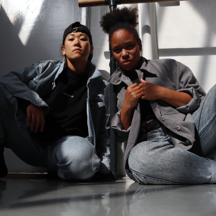 Yukiko Masui and Sarah Golding, both dressed in double denim, sitting down on the floor looking downwards into the camera.