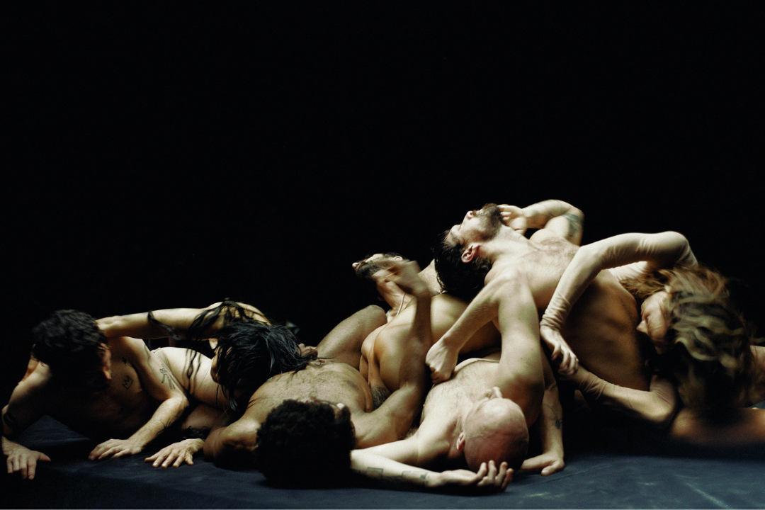 A group of people intertwined in a chaotic pose against a dark background. They're minimally dressed with various limbs and heads visible, creating a complex, entangled formation.
