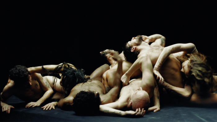 A group of people intertwined in a chaotic pose against a dark background. They're minimally dressed with various limbs and heads visible, creating a complex, entangled formation.