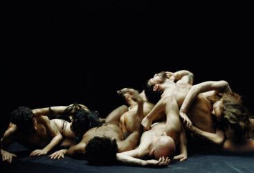 A group of people intertwined in a chaotic pose against a dark background. They're minimally dressed with various limbs and heads visible, creating a complex, entangled formation.