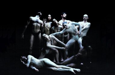 A group of minimally dressed individuals pose together, showcasing their bodies against the dark background.