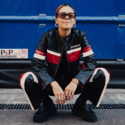 A woman wearing sunglasses and a black and red leather jacket with matching trousers, is sat crouching down on her knees in the street, in front of a blue background.