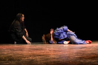 A performer in a blue outfit with red shoes is dramatically posed on the stage floor, while another figure kneels nearby in a black outfit.