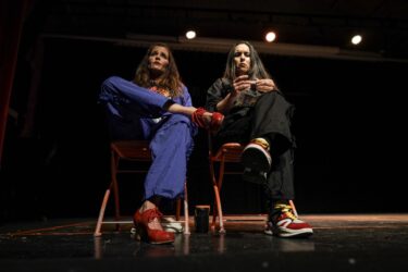 Two people sit on wooden chairs on stage, one in a blue tracksuit and red heeled shoes, the other in a black tracksuit and colorful trainers. The woman on the right is rolling a cigarette.