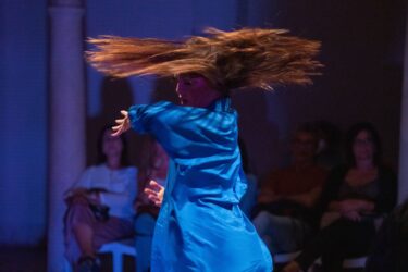 A dancer in a vibrant blue outfit spins dramatically, her hair sweeping around her, with an audience watching in dim lighting.