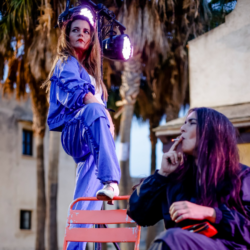 A performer in a blue tracksuit stands confidently on a red chair, with vibrant stage lights illuminating the scene and palm trees in the background. In the foreground, another person is smoking a cigarette.