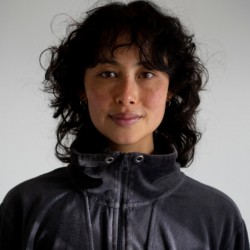 A person with short dark curly hair is wearing a dark grey zip-up jacket, standing against a neutral background.