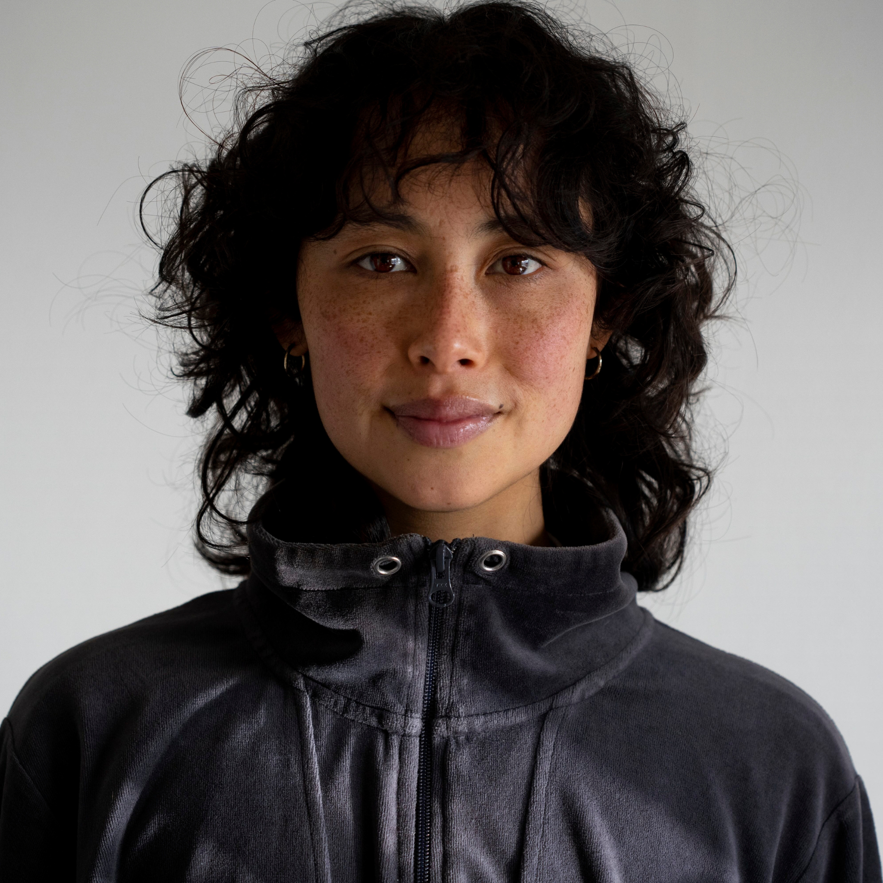 A person with short dark curly hair is wearing a dark grey zip-up jacket, standing against a neutral background.