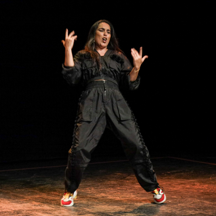 A singer performs energetically on stage, wearing a black tracksuit and vibrant trainers, against a dark backdrop.