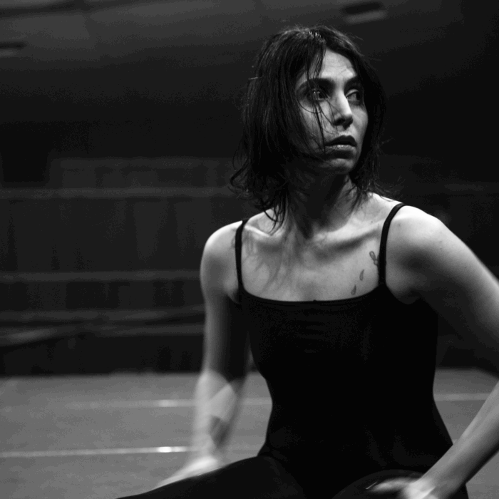 A performer with damp, tousled hair, wearing a black leotard and tights sits on stage with legs outstretched in front of her, looking inquiringly to the side.