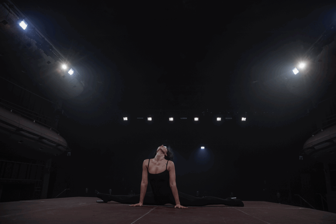 A performer in a black leotard performs the splits on stage, illuminated by bright overhead lights in a dimly lit theatre.