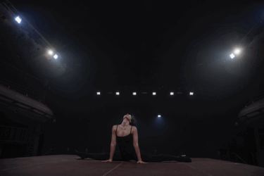 A performer in a black leotard performs the splits on stage, illuminated by bright overhead lights in a dimly lit theatre.