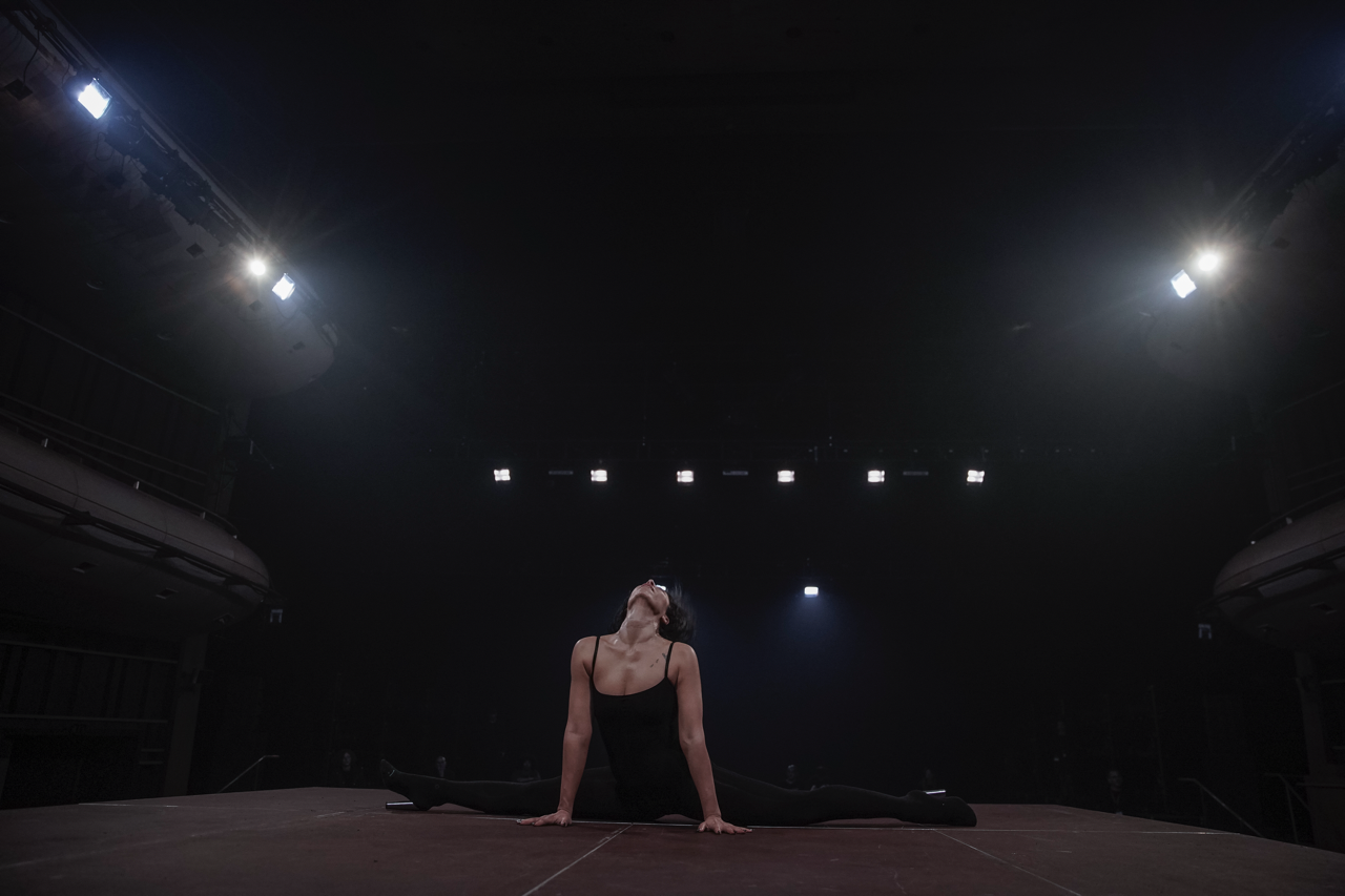 A performer in a black leotard performs the splits on stage, illuminated by bright overhead lights in a dimly lit theatre.