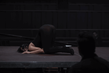 A performer in a black leotard assumes a dramatic pose on stage, showcasing flexibility and artistry in a dimly lit setting.