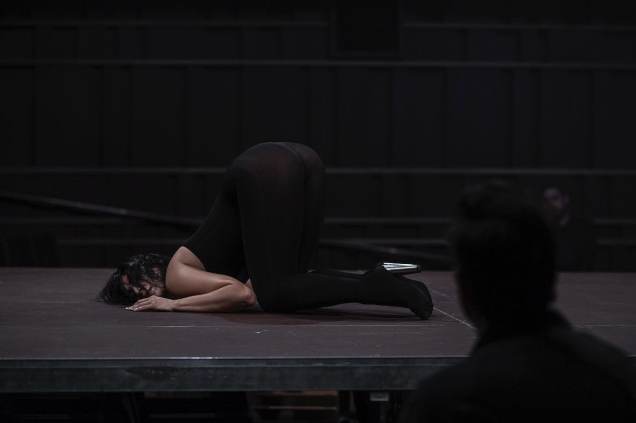 A performer in a black leotard assumes a dramatic pose on stage, showcasing flexibility and artistry in a dimly lit setting.
