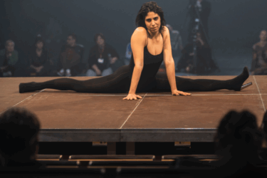 A performer in a black leotard performs the splits on stage, illuminated by bright overhead lights in a dimly lit theatre.