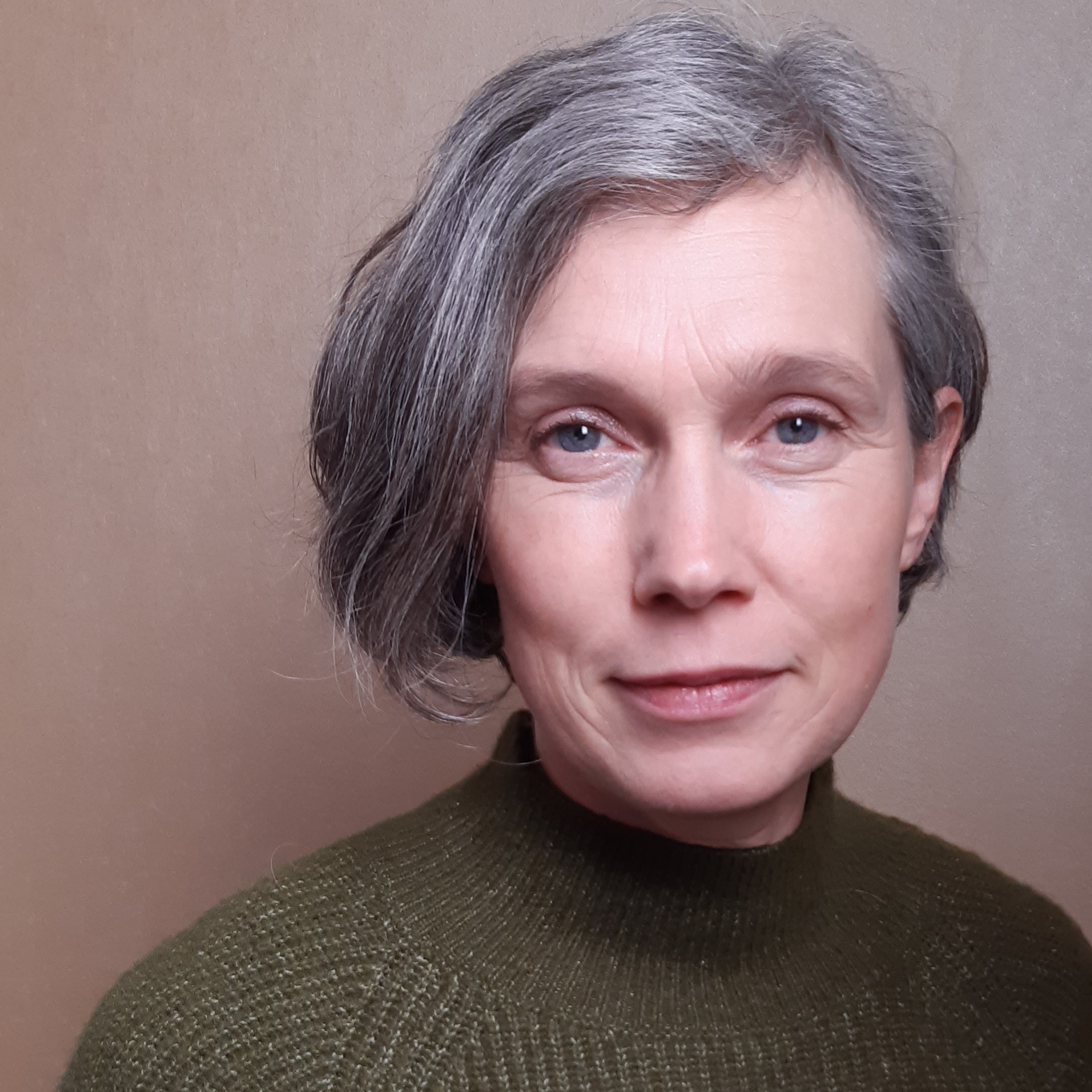 A portrait of a middle-aged woman. She has dark hair graying at the front. The woman is smiling gently and looking directly at the camera. She is wearing a green high-neck wool sweater